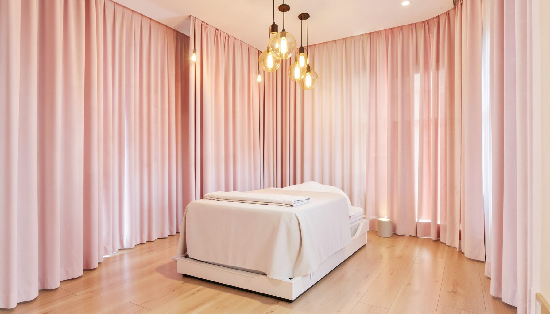 Không gian spa thanh lịch với rèm vải mềm màu hồng nhạt, đèn thả trần ánh sáng vàng ấm và sàn gỗ sáng màu
